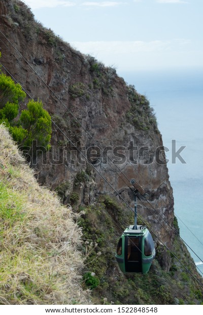 Green cable car (locally:\
Teleferico) cabin that hovers over the emptiness next to vertical\
cliff near town Achadas Da Cruz. North of Madeira island,\
Portugal.