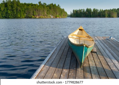 Green Canoe Rest On A Lake Wooden Pier On A Sunny Summer Day At The Cottage. A Wood Pier Is Visible Across The Water.