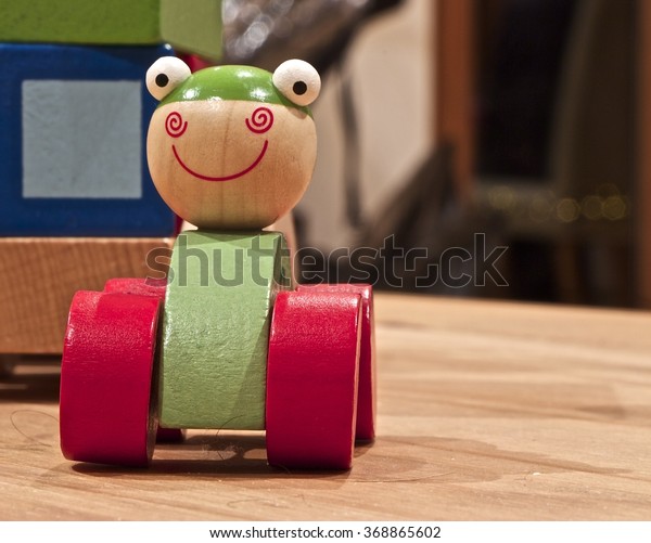 Green car toy with big red wheels on a wooden\
table, a big smile and wide white\
eyes