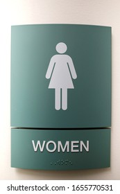 A Green Colored Curved Women's Bathroom Sign Is Hanging On The Wall, With Women Writted In Braile.