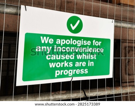 Green construction sign attached to temporary fencing apologising for inconvenience during building works in Manchester, UK.