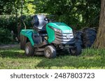 Green contemporary lawnmower on four wheels, located in shade of tree on ground. New turquoise sedentary machine designed to take care of yard near residental house, maintain right level of grass