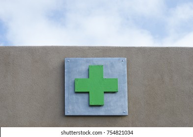 A Green Cross, The Symbol Used For Medical Marijuana, Is Shown On The Exterior Of A Stucco Building, With Sky Above - Plenty Of Space For Your Own Message.