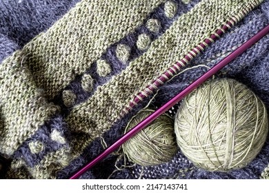 Green and dark grey wool sweater in the process of being knitted. Photograph shows textured knitting including stripes and bobbles, knitting needles and balls of yarn.