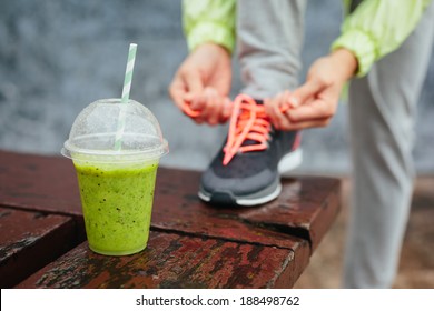 Green detox smoothie cup and woman lacing running shoes before workout on rainy day. Fitness and healthy lifestyle concept.