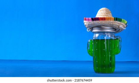 Green Drink In Cactus Shaped Glass Jar With Sombrero Hat On Blue Background. Refreshing Summer Beverage. Mexican Vacation Concept. Mockup With Copy Space