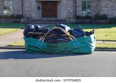 Green dumpster bag full of black plastic garbage bags on an asphalt street near the curb in front of a large house with a big green lawn