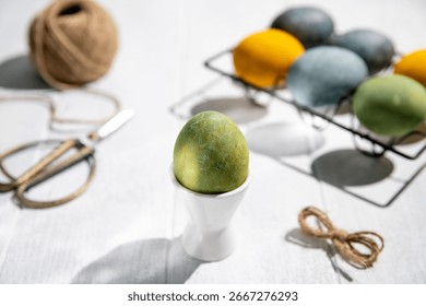 Green easter egg standing on a white egg cup on a white wooden table with other colorful eggs in a black metal egg holder, twine, and scissors, creating a festive easter scene