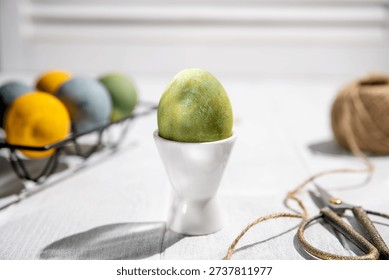 Green easter egg standing on a white egg cup on a white wooden table with other colorful eggs in a black metal egg holder, twine, and scissors, creating a festive easter scene