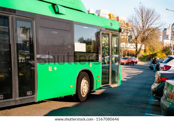 Green electric bus moves along the city\'s street\
near the parked cars. Zero emission. Public transport. Urban.\
E-bus. Ecology