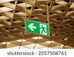 A green emergency exit sign hangs from the ceiling of a building. The sign displays a left arrow and a running figure, indicating the direction of an exit route.