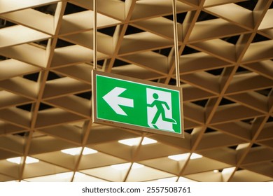 A green emergency exit sign hangs from the ceiling of a building. The sign displays a left arrow and a running figure, indicating the direction of an exit route.