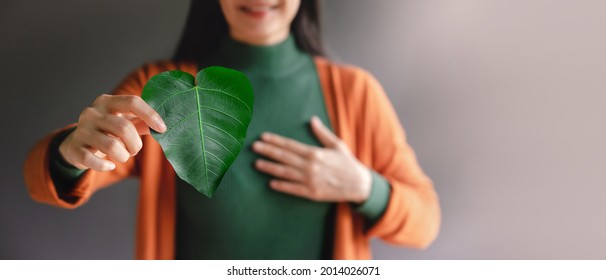 Green Energy, ESG, Renewable And Sustainable Resources. Environmental And Ecology Care Concept. Close Up Of Smiling Woman Holding A Heart Shape Green Leaf, Presenting To Camera
