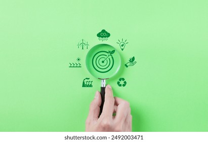 Green Energy and Sustainability Concept. Hand holding a magnifying glass focusing on a target with a leaf, surrounded by green energy and sustainability icons on a green background. Carbon footprint,