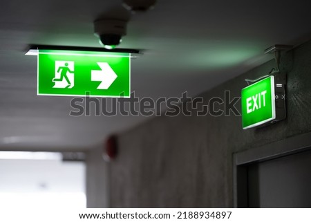 A green fire exit sign is placed on the ceiling along the dimly lit corridor and there is green exit sign on the exit door.