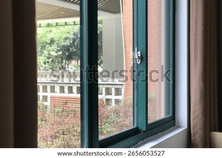 A green - framed double - hung window, partially open, reveals a view of a campus - like outdoor scene with brick buildings, white railings, and lush greenery, blending indoor calm with nature.