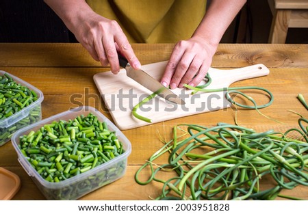 Green garlic arrows are cut with a knife on a wooden board. Preparation of food supplies for the winter. Women's hands cut the arrows of garlic.