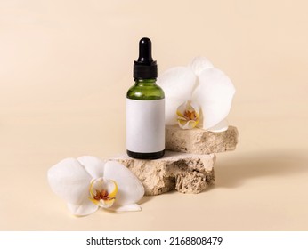 Green glass dropper bottle on stone near white orchid flowers on light yellow, close up, mockup. Skincare handmade beauty product, serum or lotion. Exotic natural cosmetics