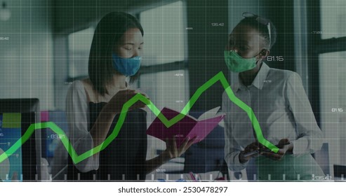 Green graph moving over two diverse female office colleagues wearing face masks discussing at office. Business data and analytics concept - Powered by Shutterstock