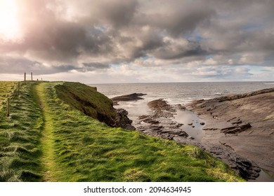 Green grass fields by the ocean and small foot path and rough stone coastline, low cloudy sky. West coast of Ireland. Irish landscape. Nobody. Nature scene