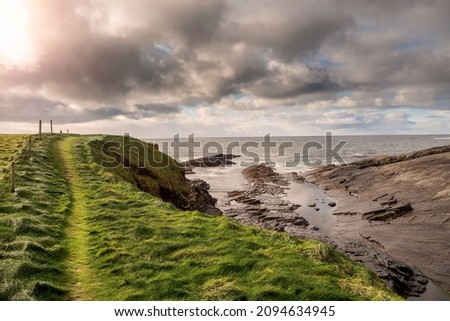 Green grass fields by the ocean and small foot path and rough stone coastline, low cloudy sky. West coast of Ireland. Irish landscape. Nobody. Nature scene