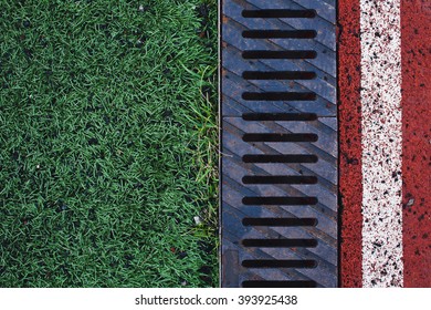 Green Grass, Rusty Drainage Rain Pipe And Red Race Track With White Line On A Stadium Overhead View. Background With Copy Space