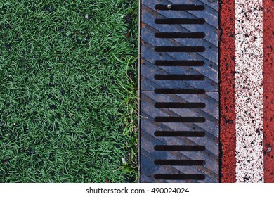 Green Grass, Rusty Drainage Rain Pipe And Red Race Track With White Line On A Stadium Overhead View. Background With Copy Space