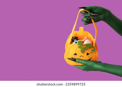 Green hands of witch with bag and candies on purple background. Halloween celebration - Shutterstock ID 2359899725