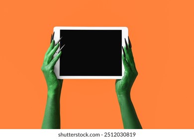 Green hands of witch with modern tablet computer on orange background. Halloween celebration - Powered by Shutterstock