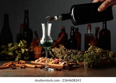 Green herbal liquor is poured from a vintage bottle into a glass. On a table dried herbs, flowers, spices, and old kitchen utensils. Concept of herbal medicine.