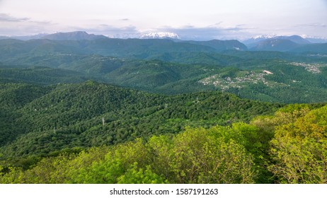 Green hills covered with dense forest and high snowy mountains in the distance. Fog and clouds on the slopes of high mountains with snowy peaks. Beautiful sunset in a hilly valley with villages. - Shutterstock ID 1587191263