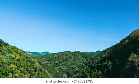 Green hills in Japan, morning, featuring a mountain landscape with trees in autumn colors green yellow orange under a clear blue sky, epitomizing serene natural beauty. SHOTLISTtravel - Powered by Shutterstock