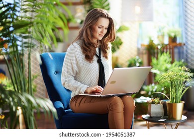 Green Home. 40 Years Old Housewife With Long Wavy Hair In The Modern Living Room In Sunny Day In Beige Pants And Blouse Using Laptop While Sitting In A Blue Armchair.