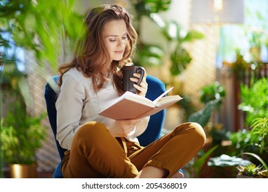 Green Home. elegant woman with long wavy hair with cup of cappuccino and book in green pants and grey blouse sitting in a blue armchair at modern home in sunny day.