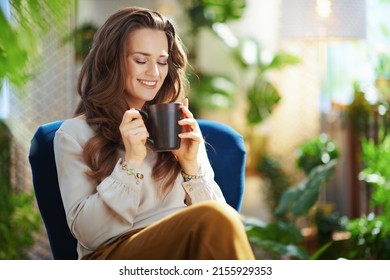 Green Home. happy middle aged woman with long wavy hair with cup of tea sitting in a blue armchair at modern home in sunny day. - Shutterstock ID 2155929353