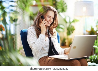 Grünes Zuhause. Happy moderne 40 Jahre alte Frau mit langen Haaren im modernen Haus an sonnigen Tagen mit einem Smartphone und Laptop beim Sitzen in einem blauen Sessel.