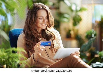 Grünes Zuhause. Hübsches, modernes Weibchen mit langen Haaren mit Kreditkarte mit Tablet PC im modernen Haus an sonnigen Tagen.