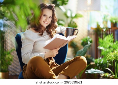 Grünes Zuhause. Happy stylische Hausfrau mit langgestrecktem Haar mit Tasse Kaffee und buchen in grüner Hose und graue Bluse sitzend in einem blauen Sessel im modernen Zuhause bei sonnigem Tag.