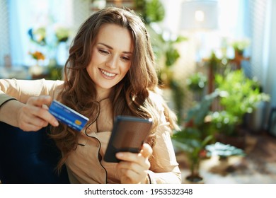 Green Home. Happy Trendy Middle Aged Woman With Long Wavy Hair With Credit Card Making Online Shopping On E-commerce Website In The Modern House In Sunny Day.