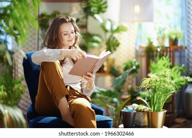 Grünes Zuhause. Entspannte, trendige Frau mittleren Alters mit langwelligen Haaren mit Buch in grünen Hosen und graue Bluse im modernen Wohnzimmer bei sonniger Nacht.