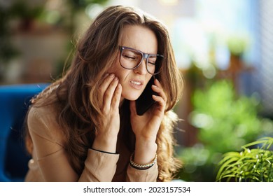 Grünes Zuhause. traurige elegante Hausfrau mittleren Alters mit langen Haaren mit Zahnschmerzen sprechen auf einem Smartphone im modernen Haus an sonnigen Tagen.