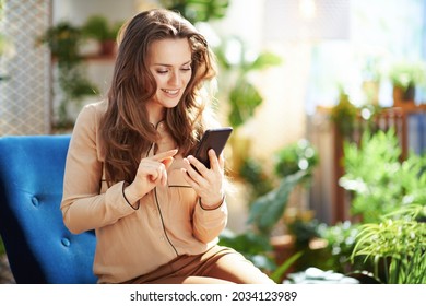 Green Home. smiling 40 years old woman with long wavy hair in the modern house in sunny day in beige pants and blouse using smartphone applications.