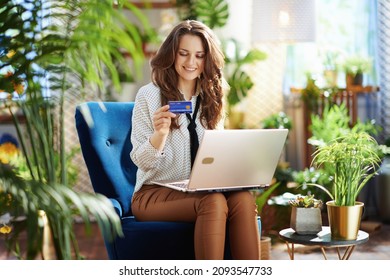 Grünes Zuhause. Lächelnde moderne 40-jährige Frau mit langweiligen Haaren mit Kreditkarte in beige Hosen und Bluse mit Laptop während sie in einem blauen Sessel im modernen Zuhause bei sonniger Nacht sitzt.