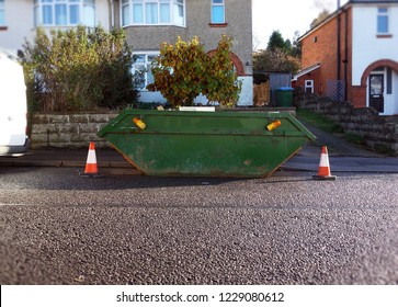 Green Industrial Rubbish Skip Bin With Orange & White Safety Cones On Side Of Street. Space To Add Text On Wet Road Surface, Driveway & Houses In Background. Renovate Building Project, House Clearance