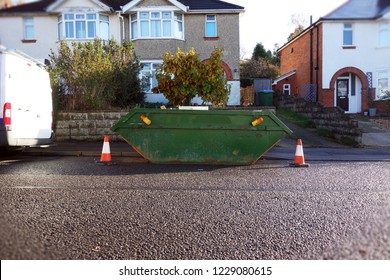 Green Industrial Rubbish Skip Bin With Orange & White Safety Cones On Side Of Street. Space To Add Text On Wet Road Surface, Driveway & Houses In Background. Renovate Building Project, House Clearance