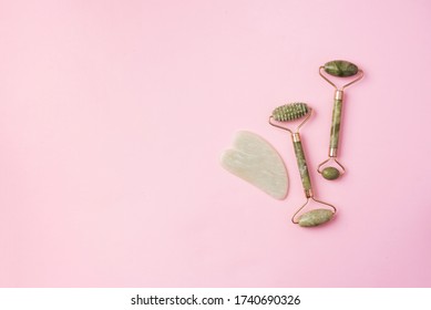 Green jade face roller and gua-sha scraper for facial acupuncture massage on pink background. Copy space. Skin care, face treatment.