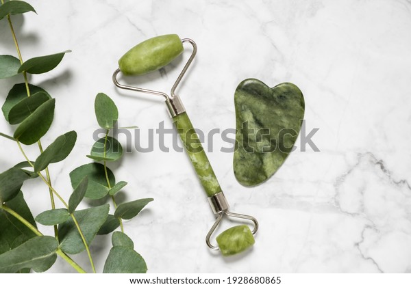 Green jade roller and gua sha stone for facial
massage and eucalyptus branch on marble background. Home beauty and
selfcare accessories. Face roller for anti age wrinkle treatment.
Top view, flat lay.
