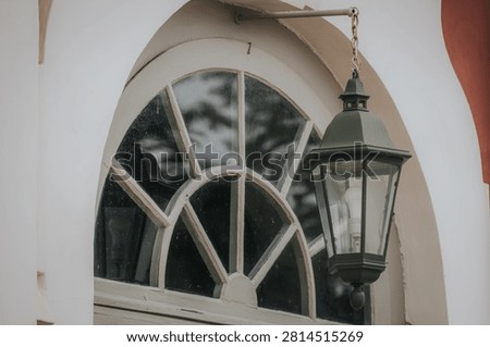 Green lantern is positioned next to an arched window with a white frame, featuring detailed glass patterns and reflections of nearby plants