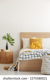 Green Leaf In Black Vase On Wooden Nightstand Next To Yellow Pillows And Blanket On White Single Wooden Bed