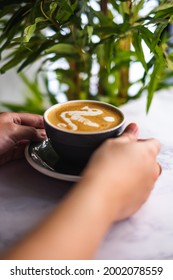 Green Leafy Background With A Close Up Photo Of Woman's Hands Holding A Black Cup Of Coffee With Seahorse Latte Art